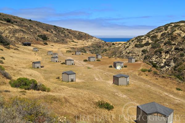 Campgrounnd wind shelters on dry, grassy terrace looking down canyon to blue ocean.