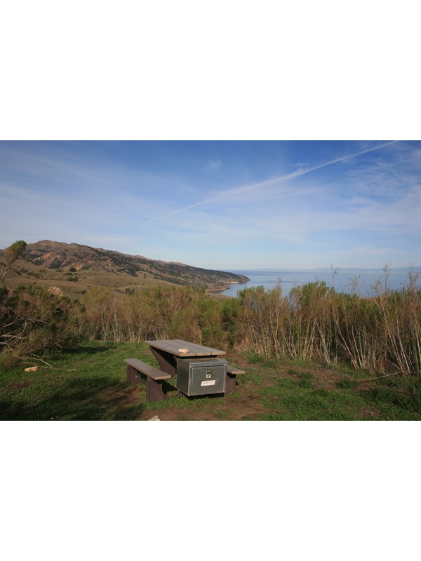 Campground picnic table on green grass overlooking ocean, coastline, and blue sky with white clouds