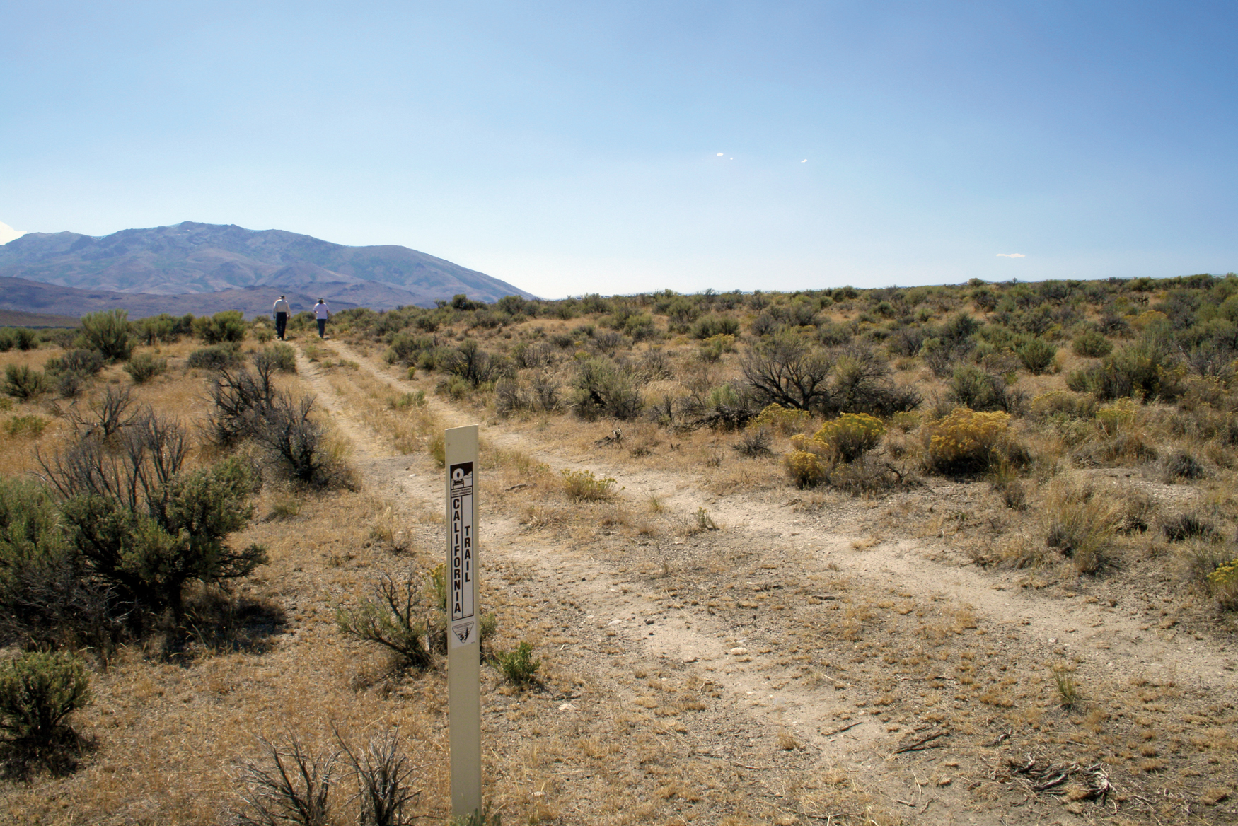 A dirt road passes through sagebrush and a white post with a mountain in the background.