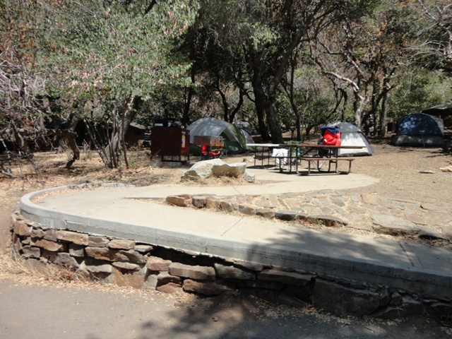 A paved ramp leads to a picnic table among oaks and tents