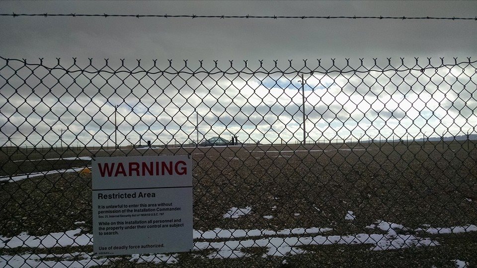 Two people are visible at the missile silo through a chain link fence