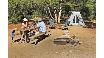 A man and a woman sit at a picnic table in the sunlight a tent and chairs surround a firepit