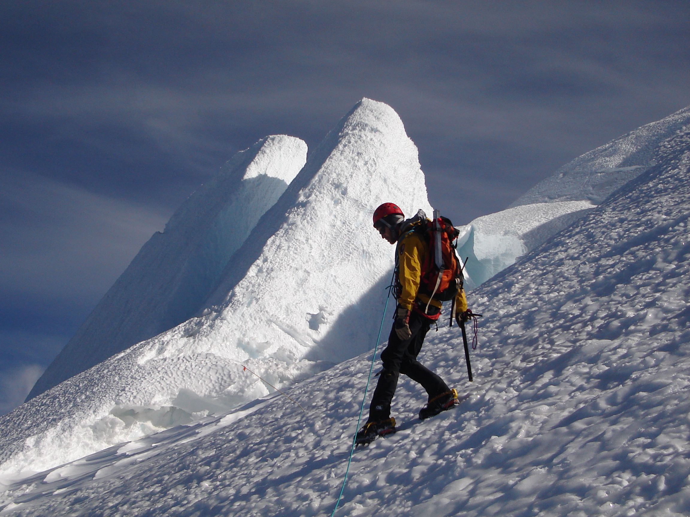Climber on glacier steps downward with icy crags in background.