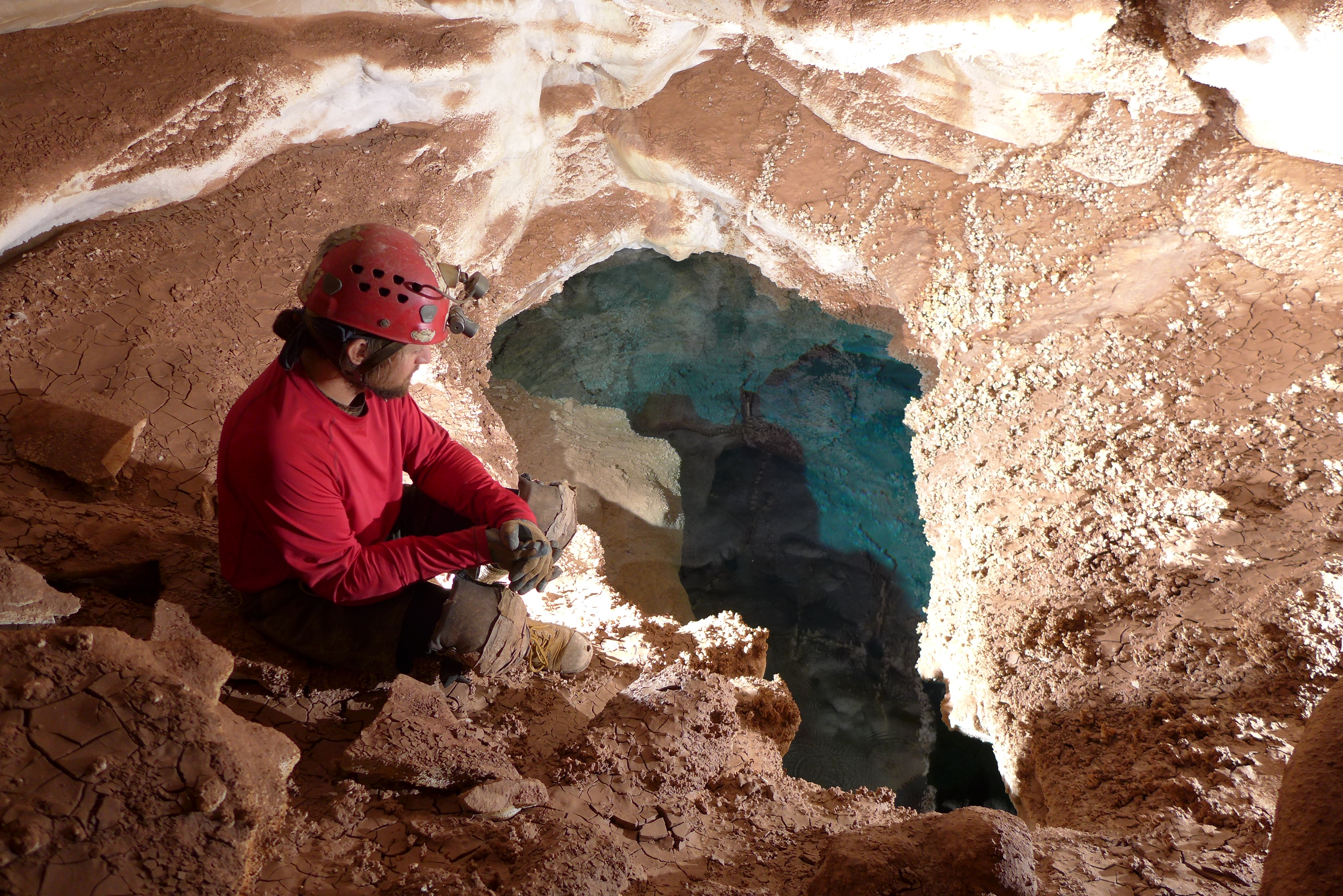 A cave explorer sits on some rocks and looks at a clear blue lake in Jewel Cave.