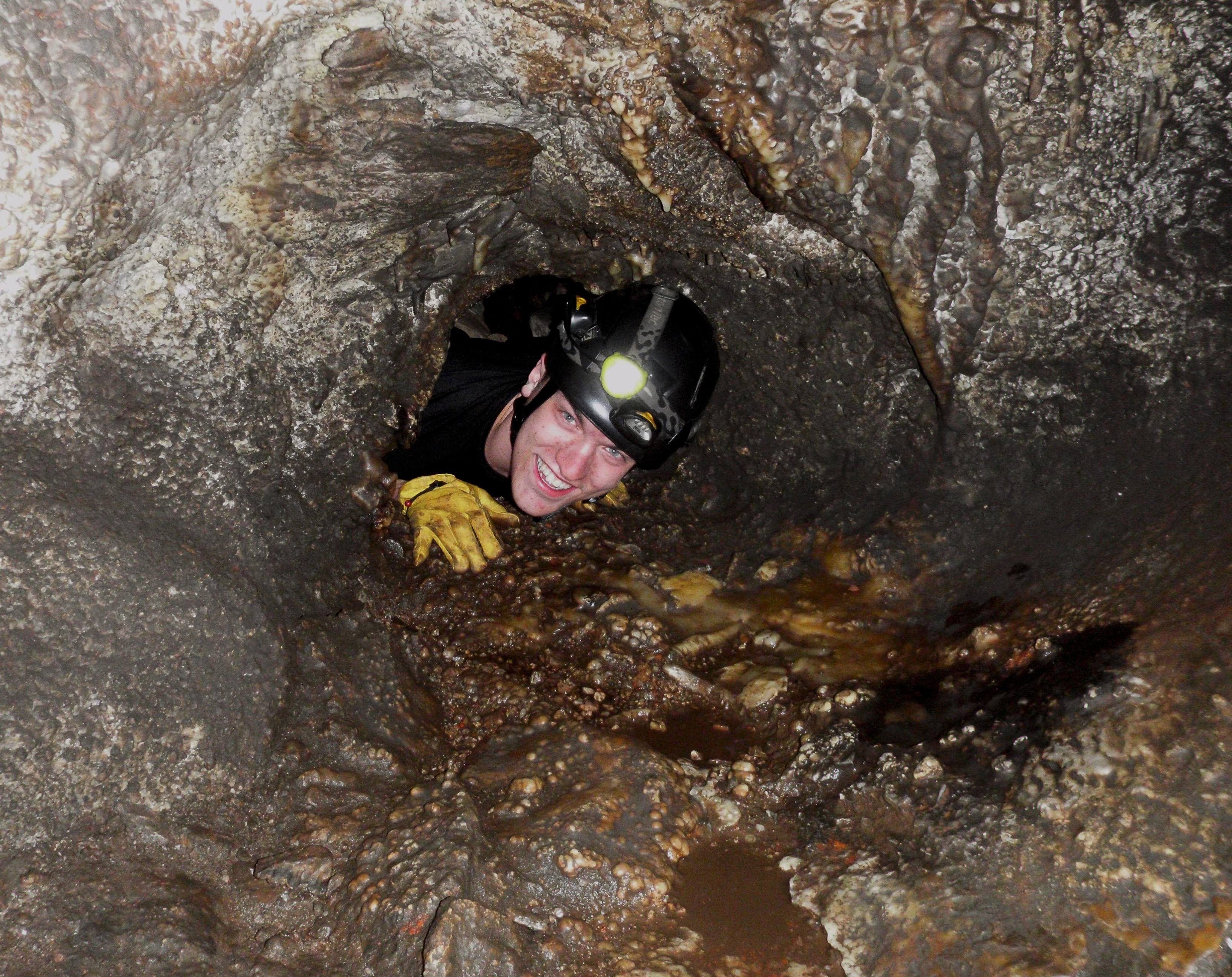 A caver squeezes through a very small and tight opening, surrounded by multi-colored rocks.