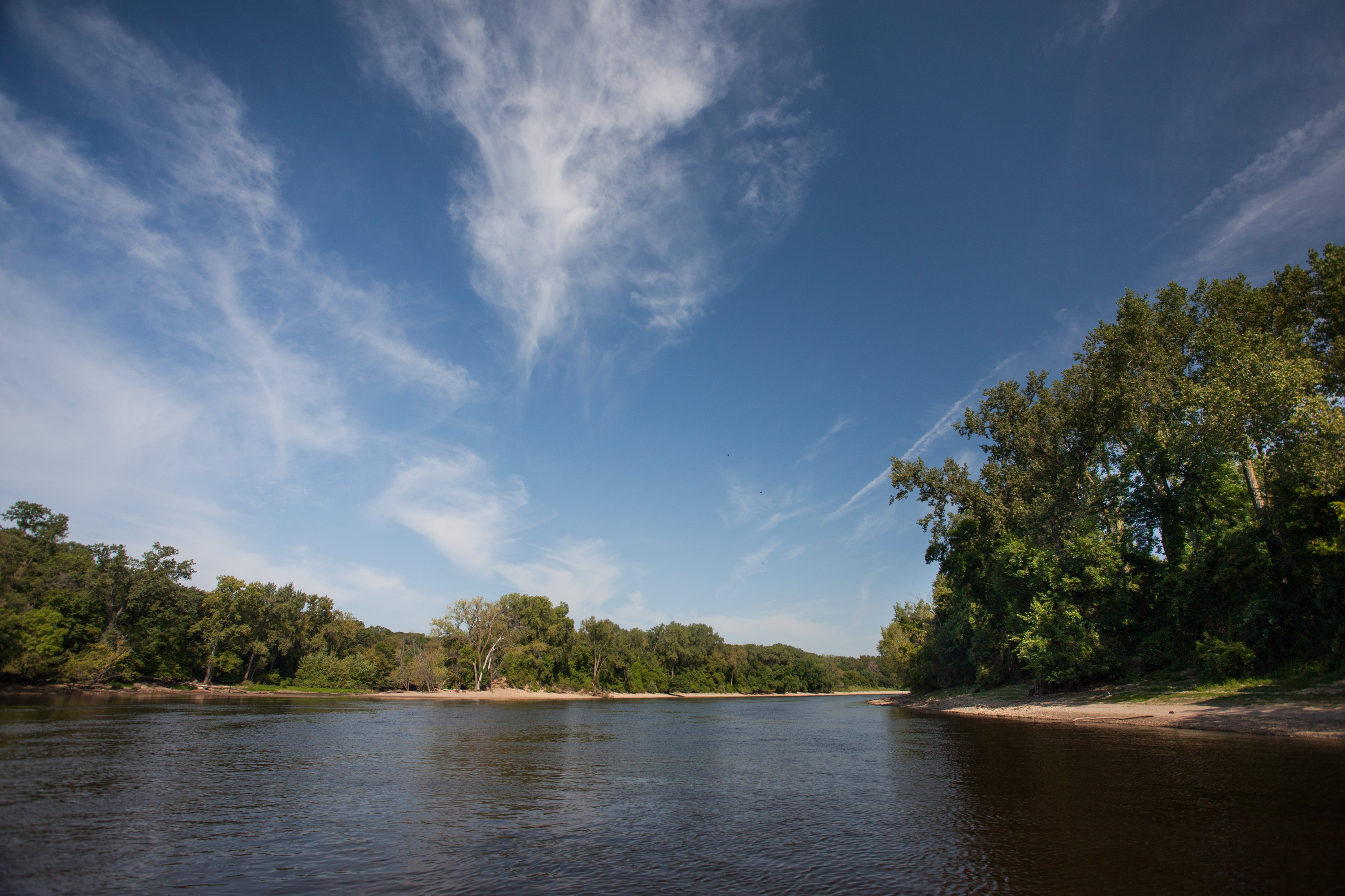 White clouds streak the blue sky over a river flanked by sandy shorelines and green trees.