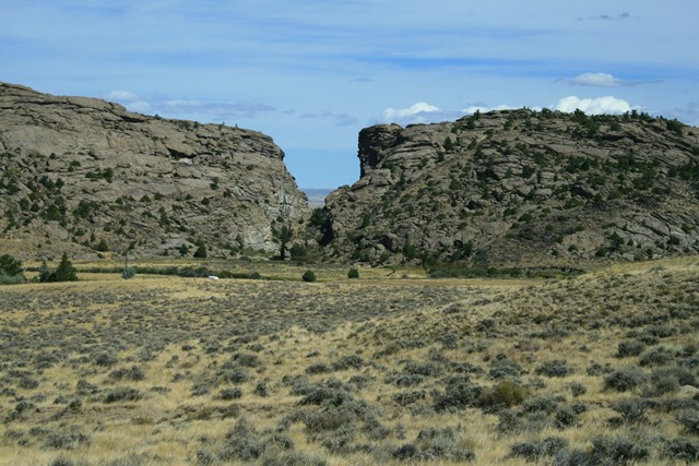 A tall rock buttress with a cleft in the center sits behind a sagebrush covered valley.