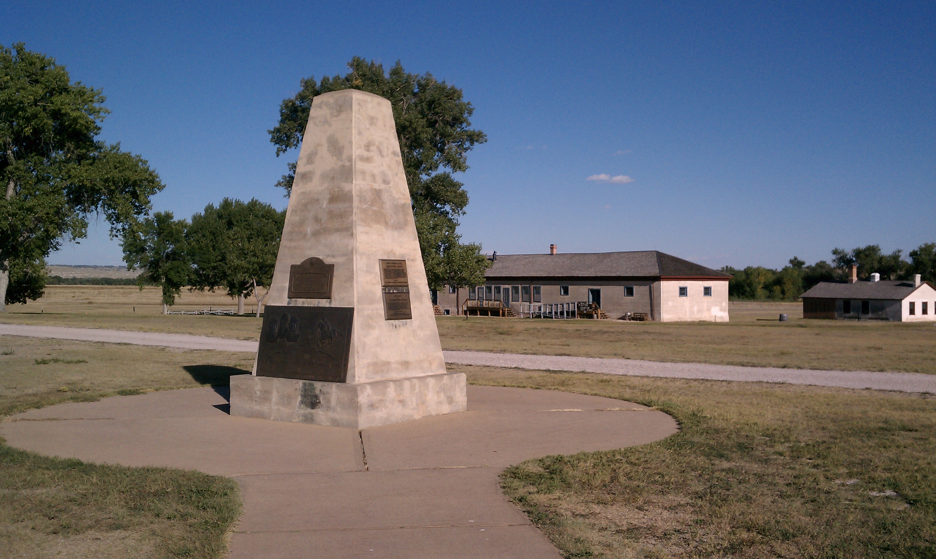 A tall obelisk monument with bronze plaques sits on a paved pathway in front of a building.