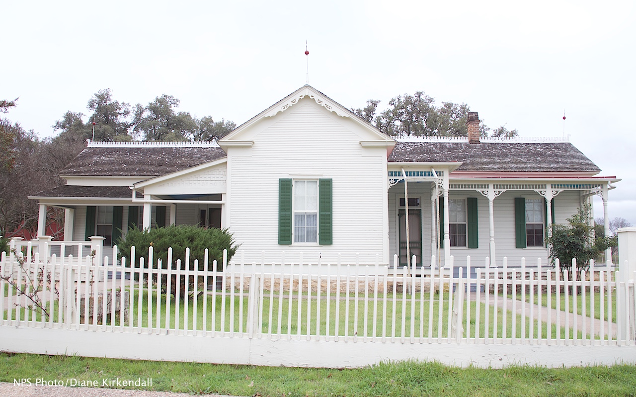 View of a one-story, white-frame house surrounded by a white picket fence.