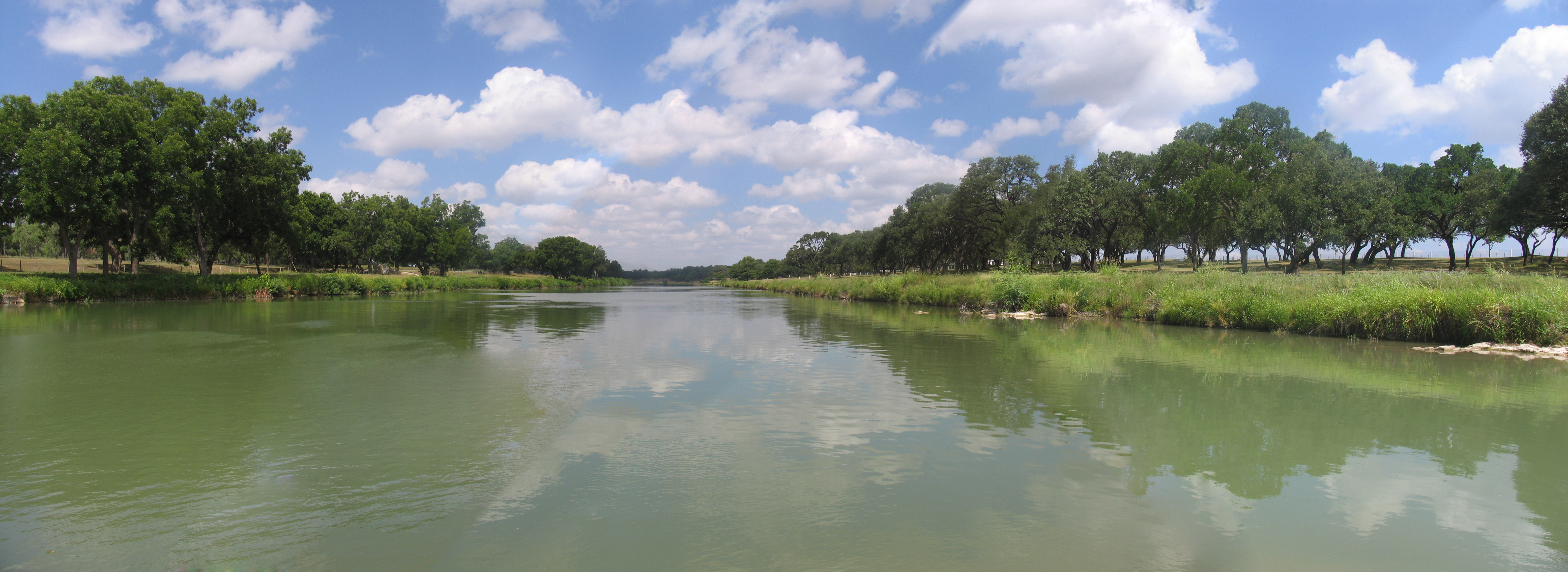 The Pedernales River with tree-lined banks under a blue sky dotted with clouds.