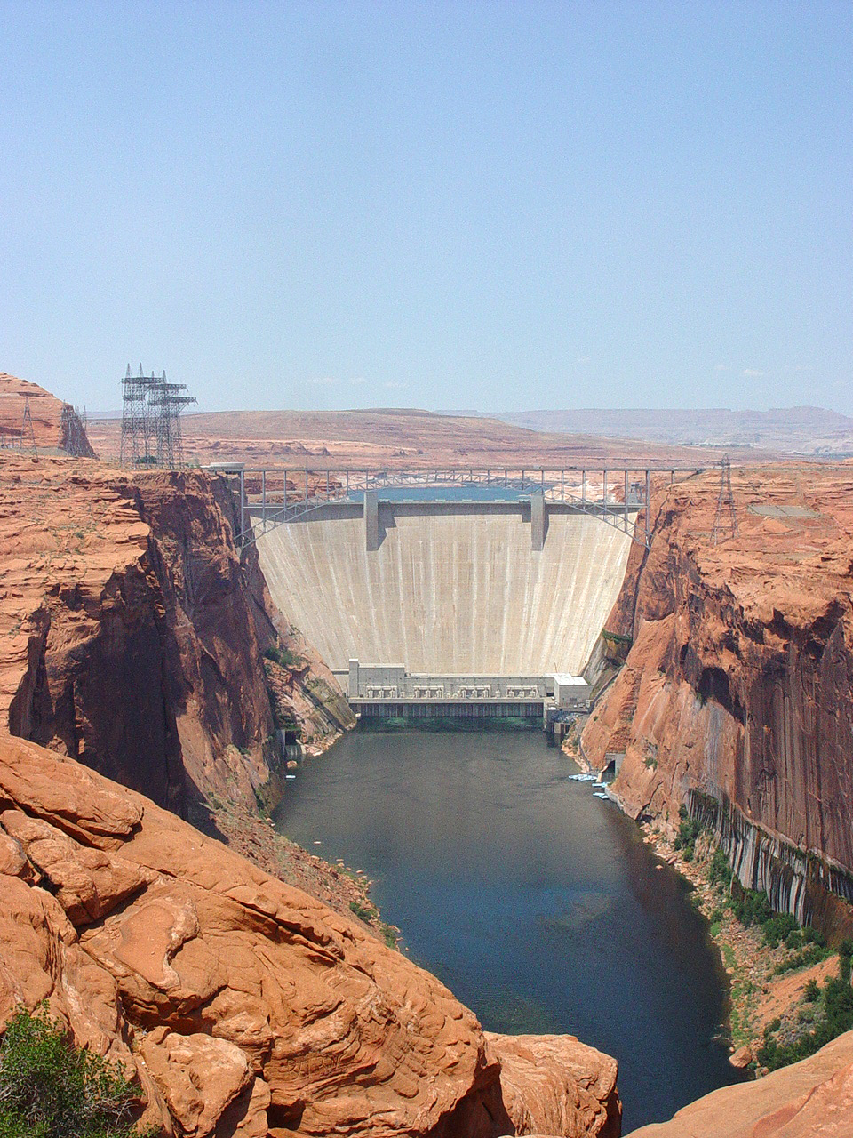 A concrete dam plugs high canyon walls. Buildings and wires on the sides.