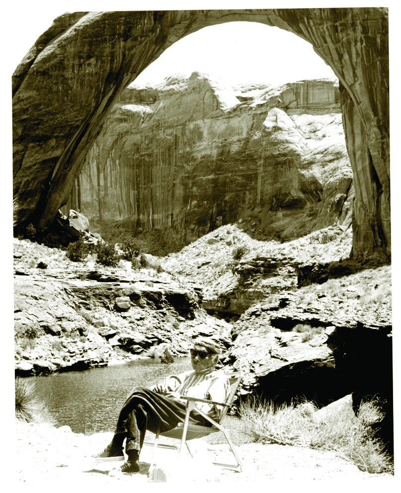 An elderly Native American man sits in a lawn chair under Rainbow Bridge