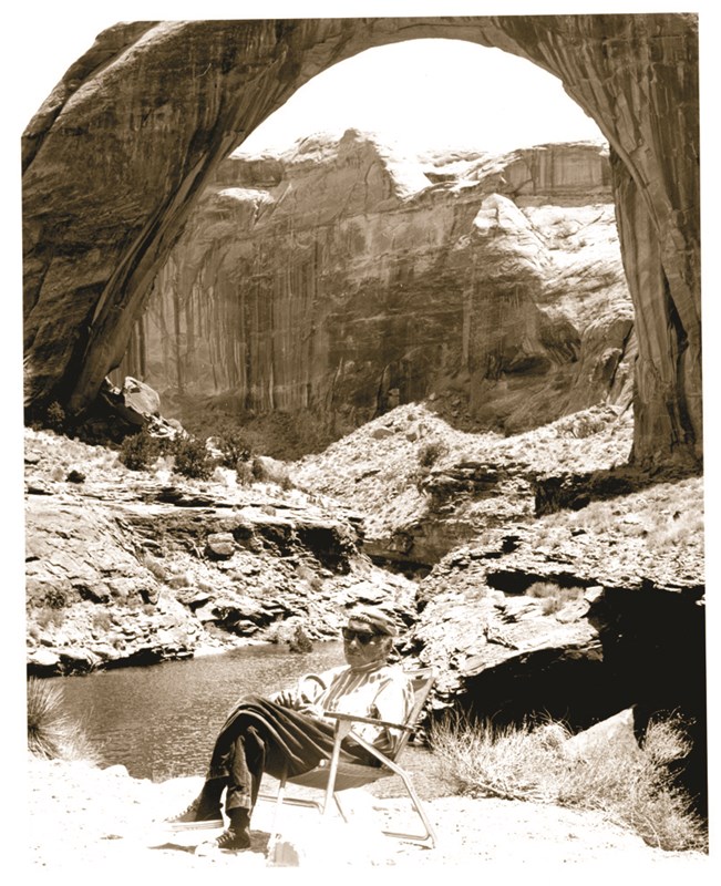 An elderly Native American man sits in a lawn chair under Rainbow Bridge