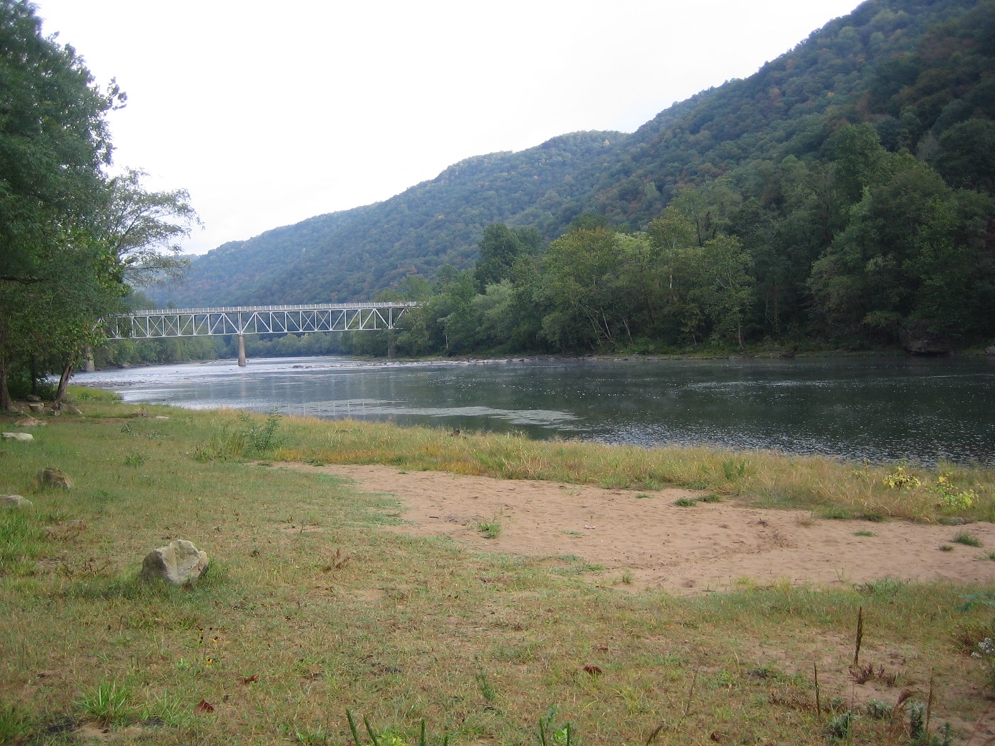 A sandy beach next to a swift flowing river with a bridge over the river in the distance
