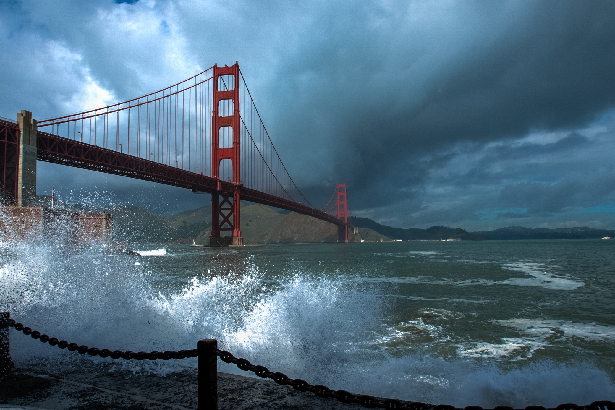 Orange Golden Gate Bridge with waves crashing in foreground and storm clouds behind.