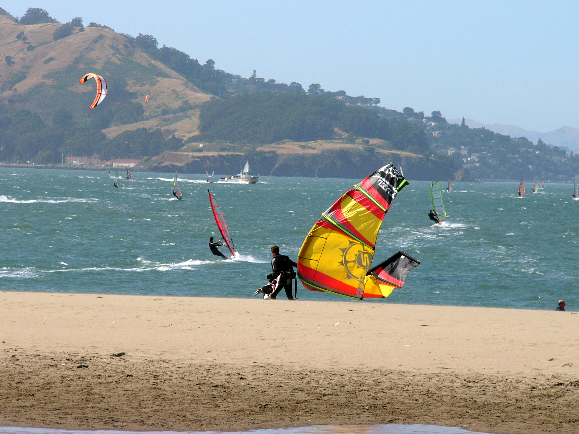A kite boarder with yellow kite on beach with wind surfers behind on the blue bay.