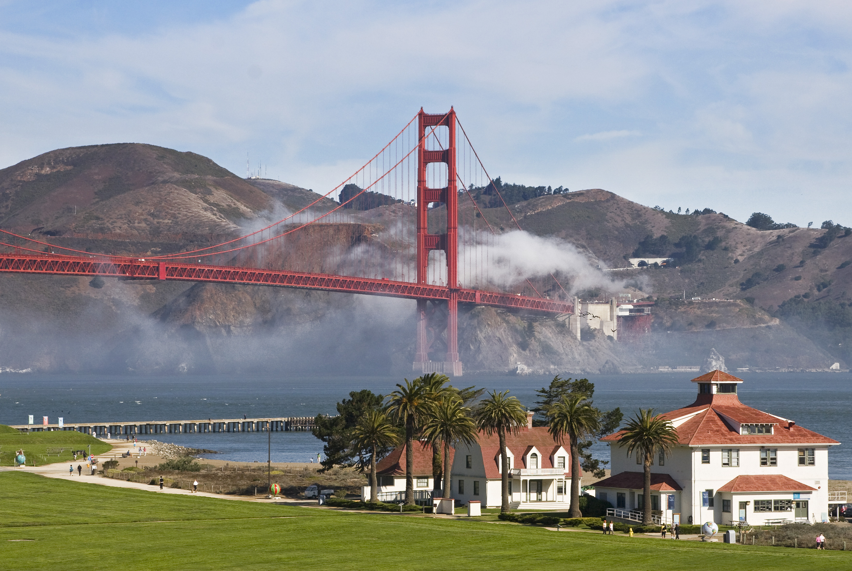 White building with red roofs on Crissy Field with blue bay and orange bridge with fog behind.