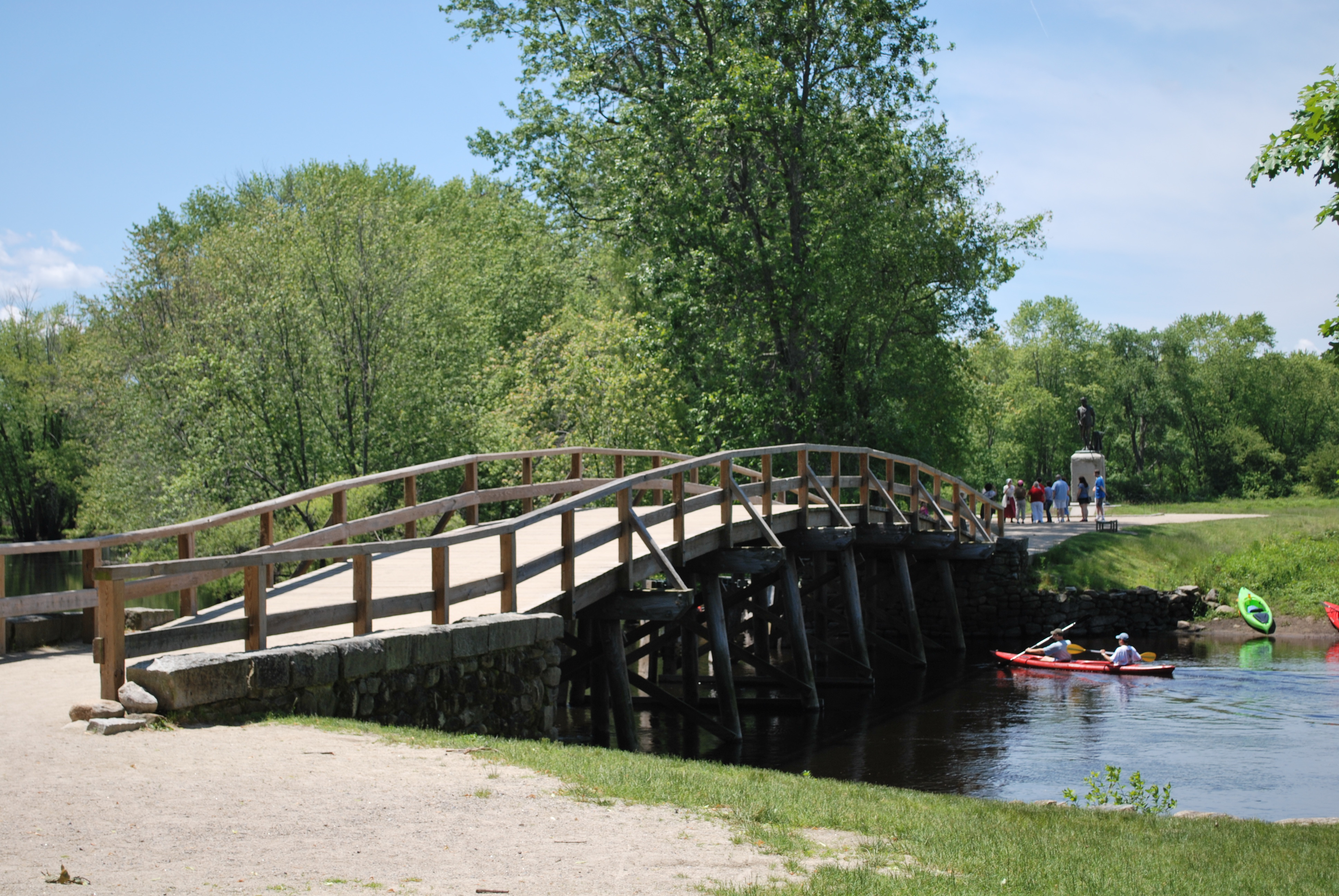 A wooden bridge spans the Concord river as people walk and canoe paddlers gluide under the bridge