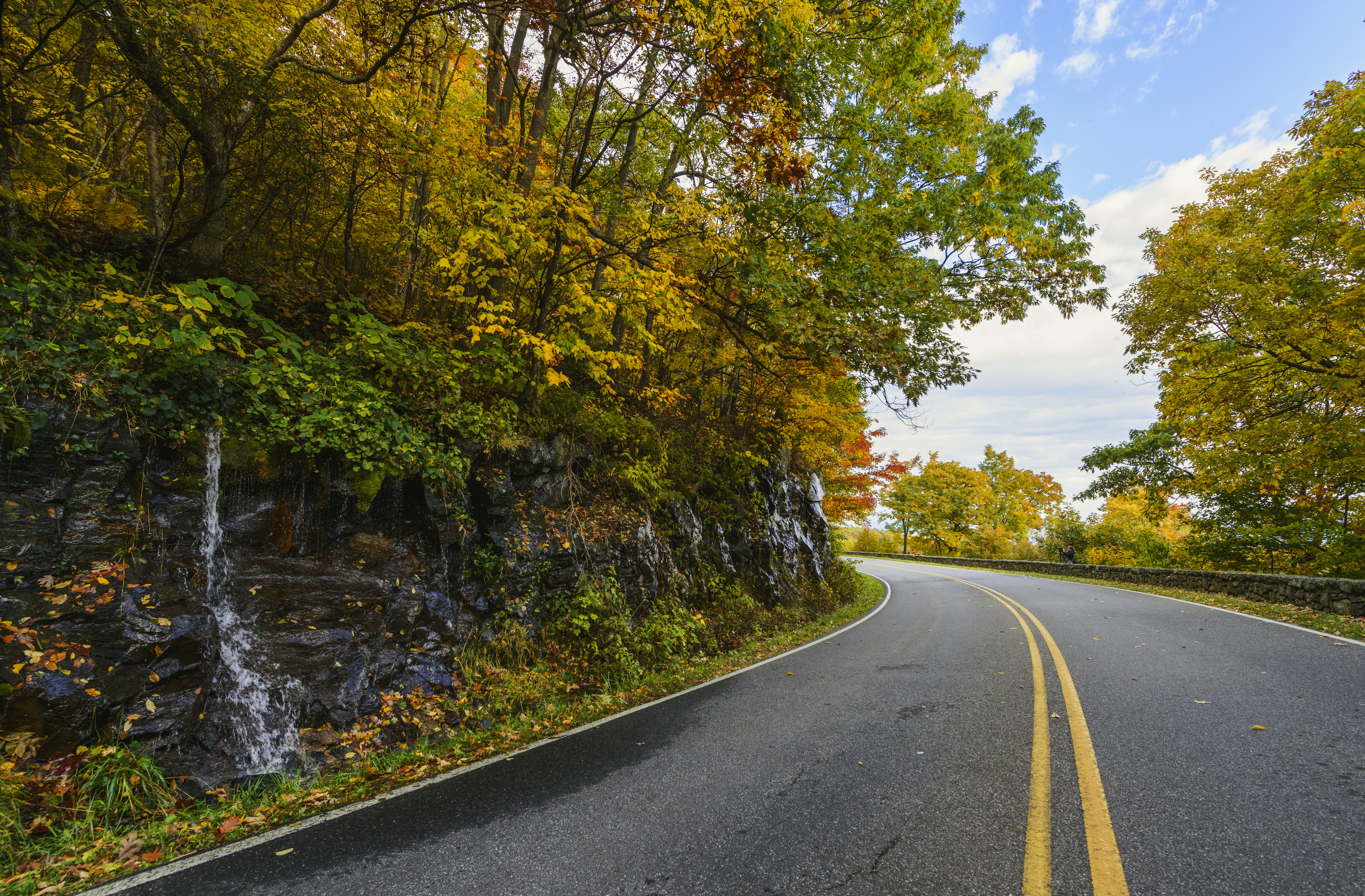 A road surrounded by fall foliage turns a curve around a small waterfall.