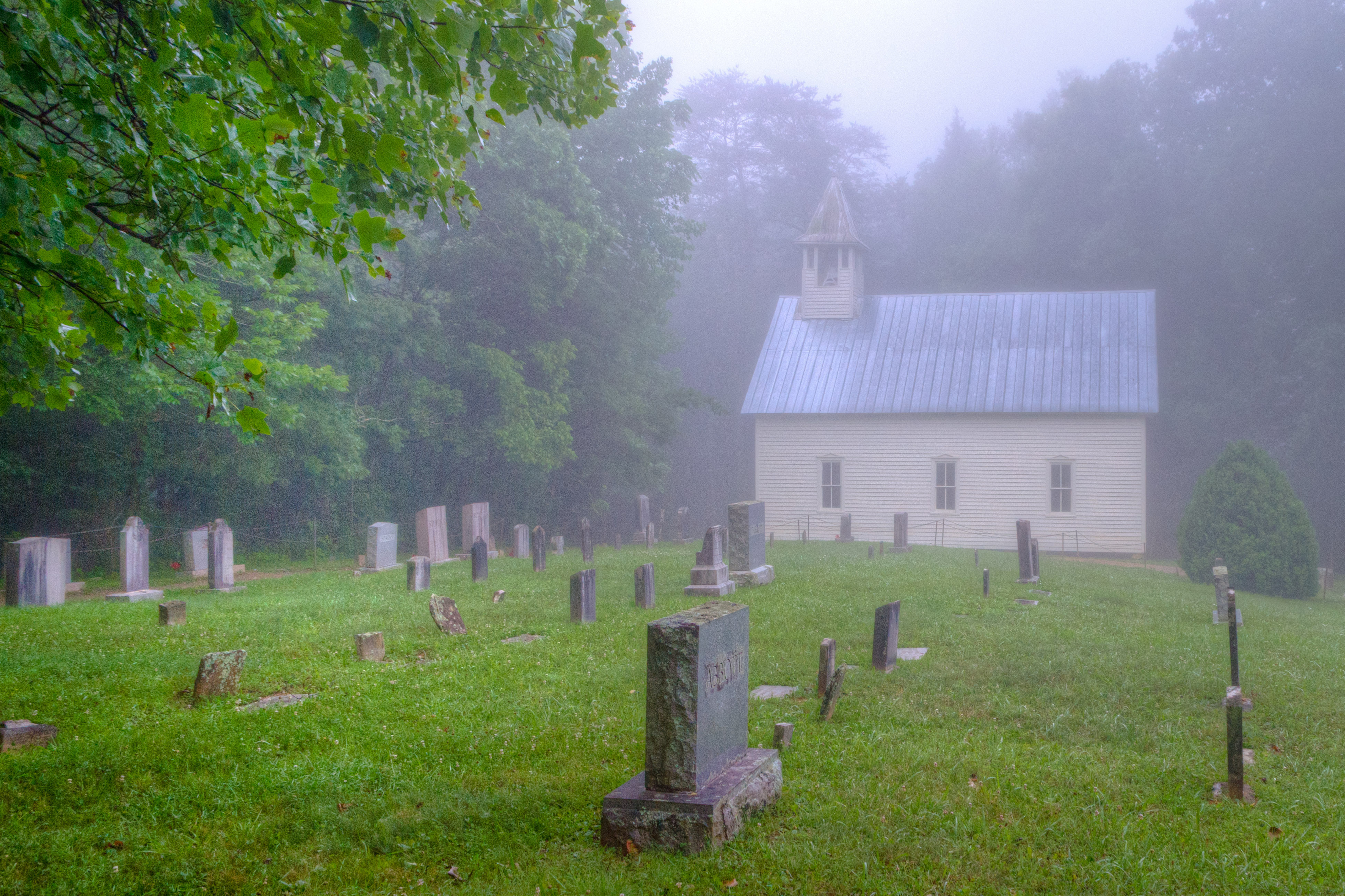 Tombstones stand in front of a white, wooden church on a foggy morning.
