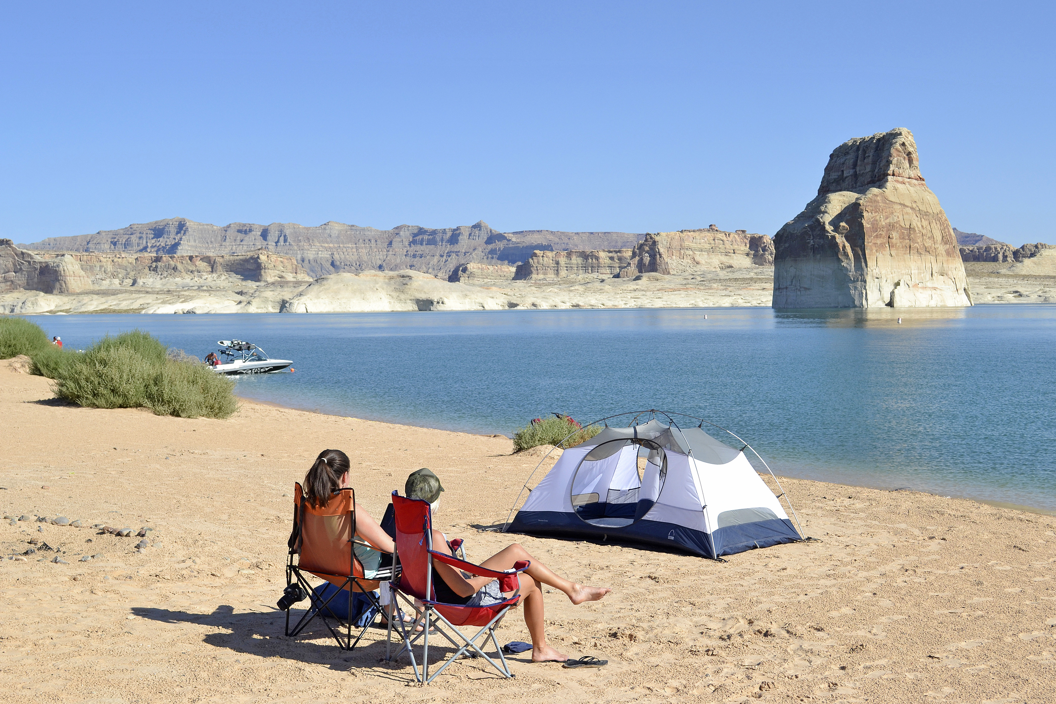 Two people sit in beach chairs in front of a tent pitched on a beach near the water's edge.