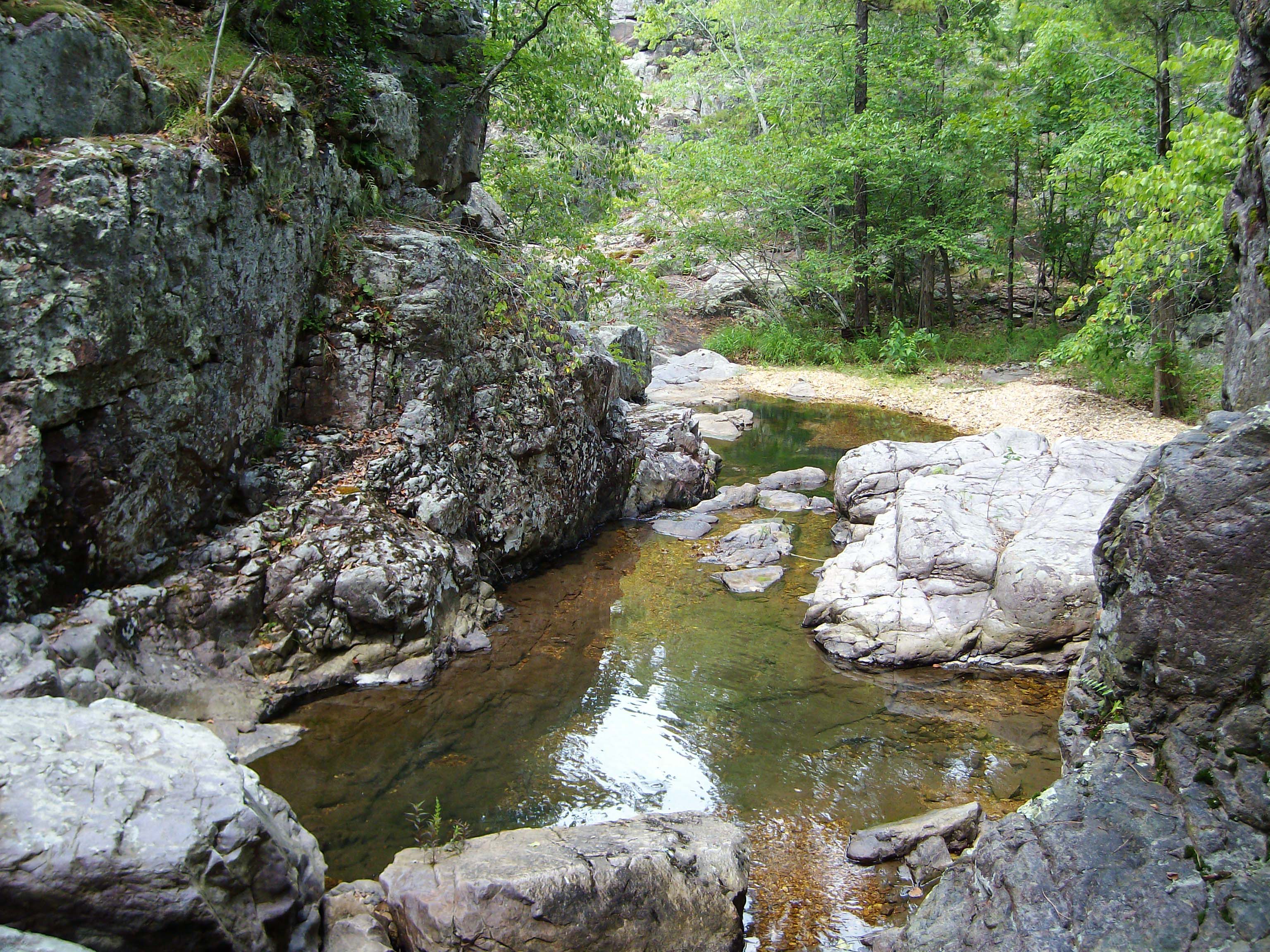 looking down through large brown/grey/purple boulders with creek in middle and forest in distance