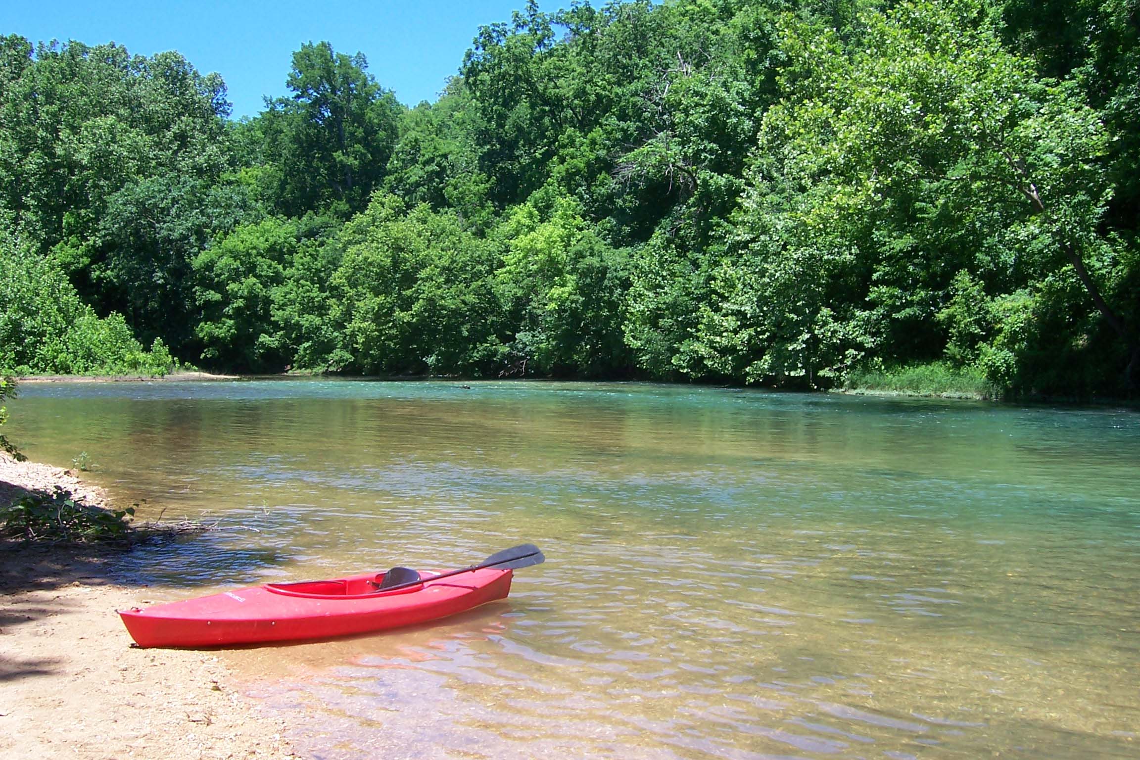 A red canoe on gravel bar with clear blue stream behind and green forest