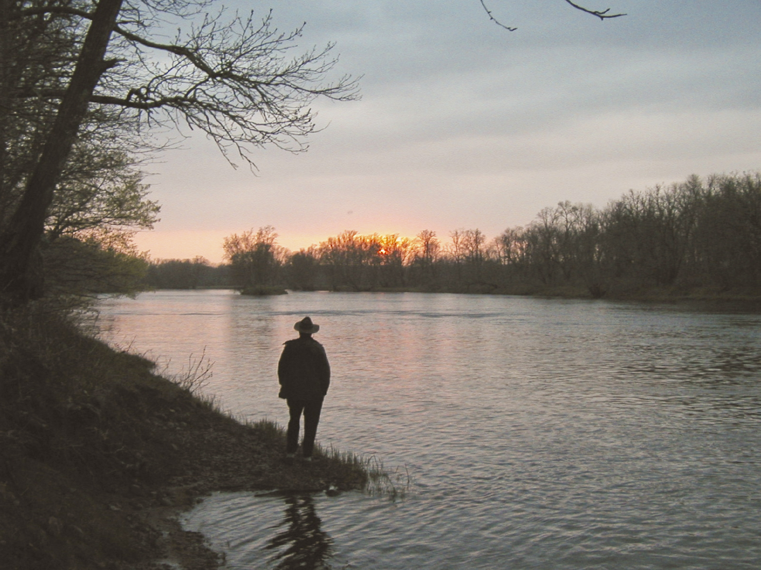 A man stands next to a river watching a sunset.