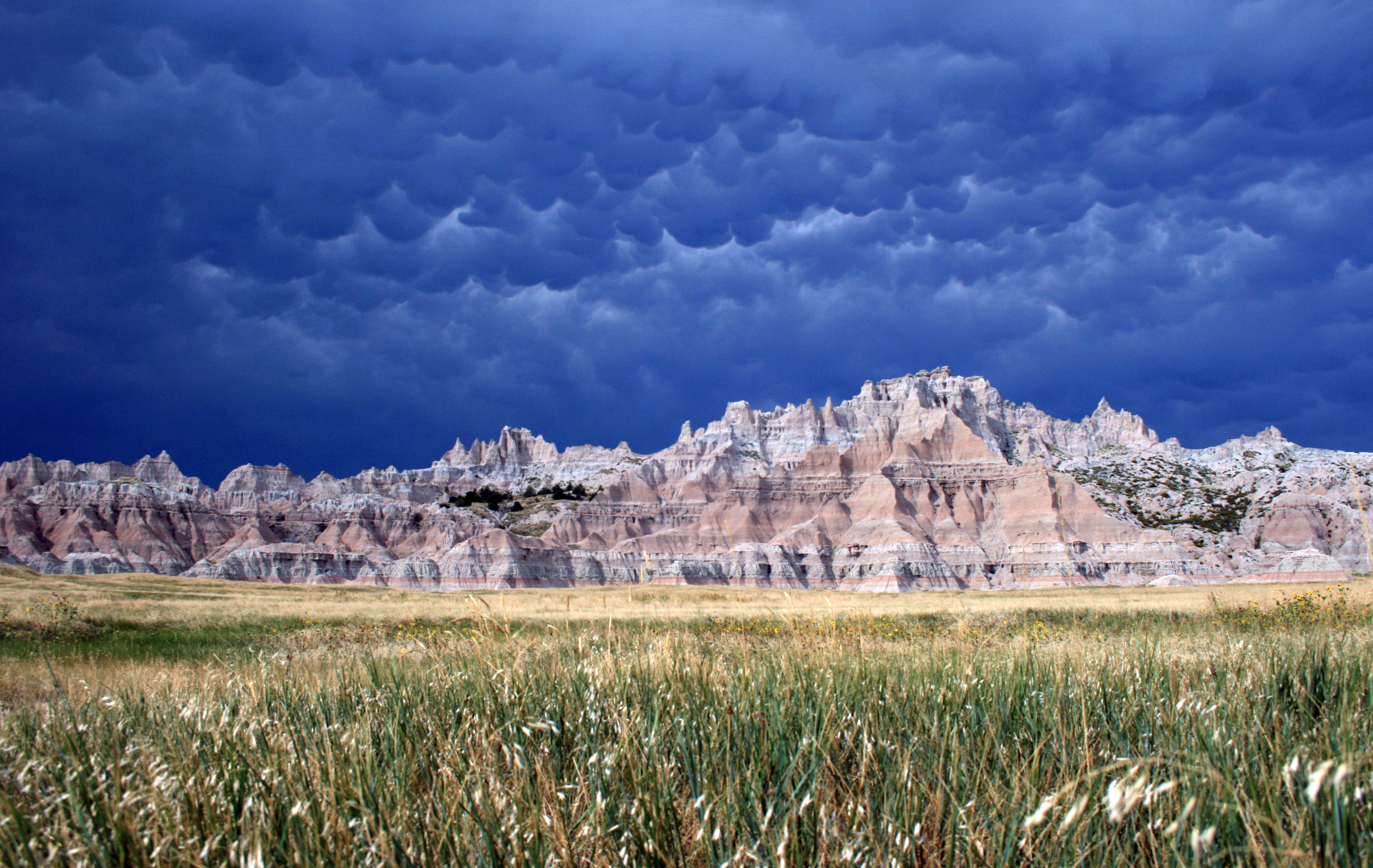 Layered badlands formations behind fields of green grass under cloudy and billowing clouds.