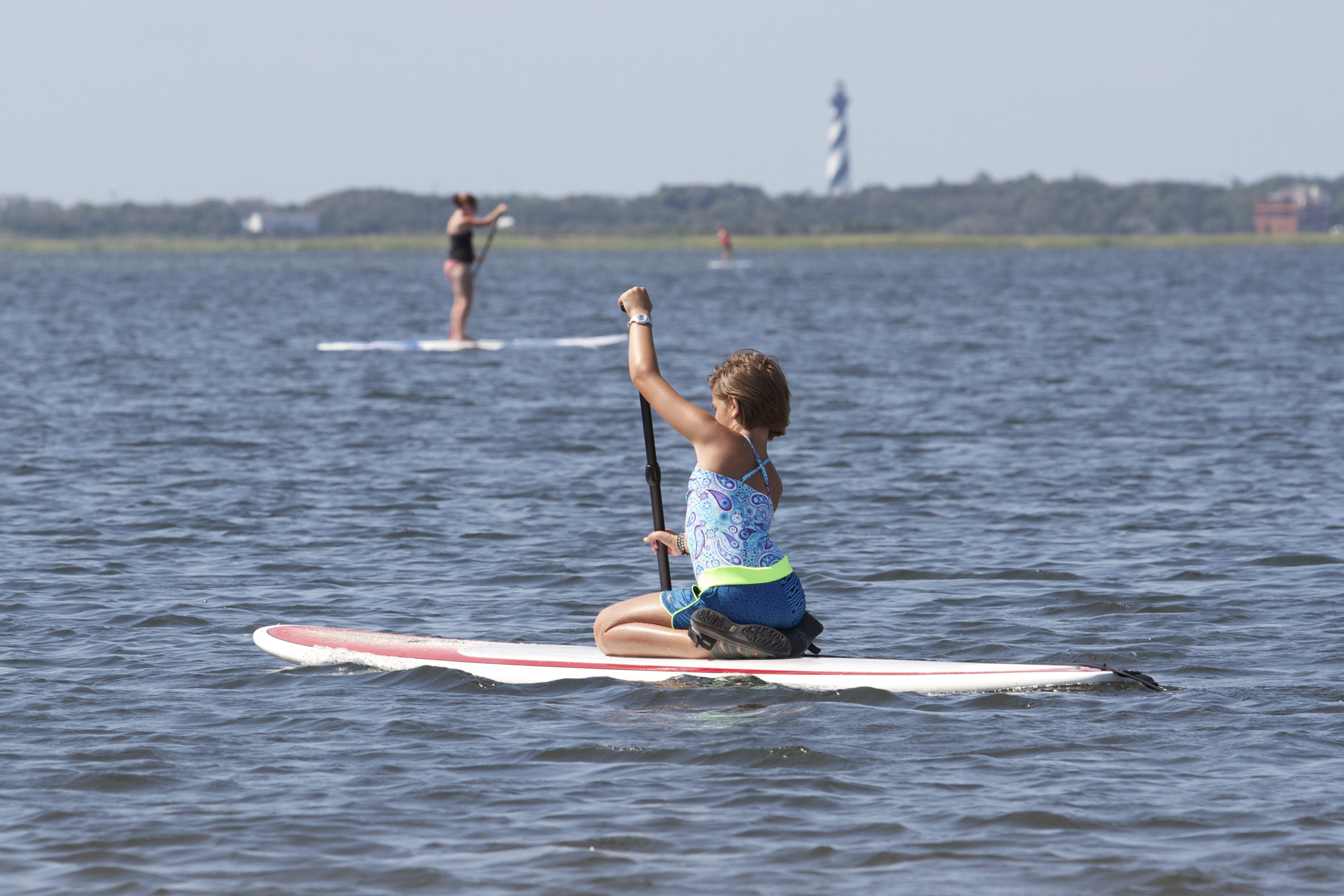 A young girl paddles across the water with the Cape Hatteras Lighthouse visible in the distance.
