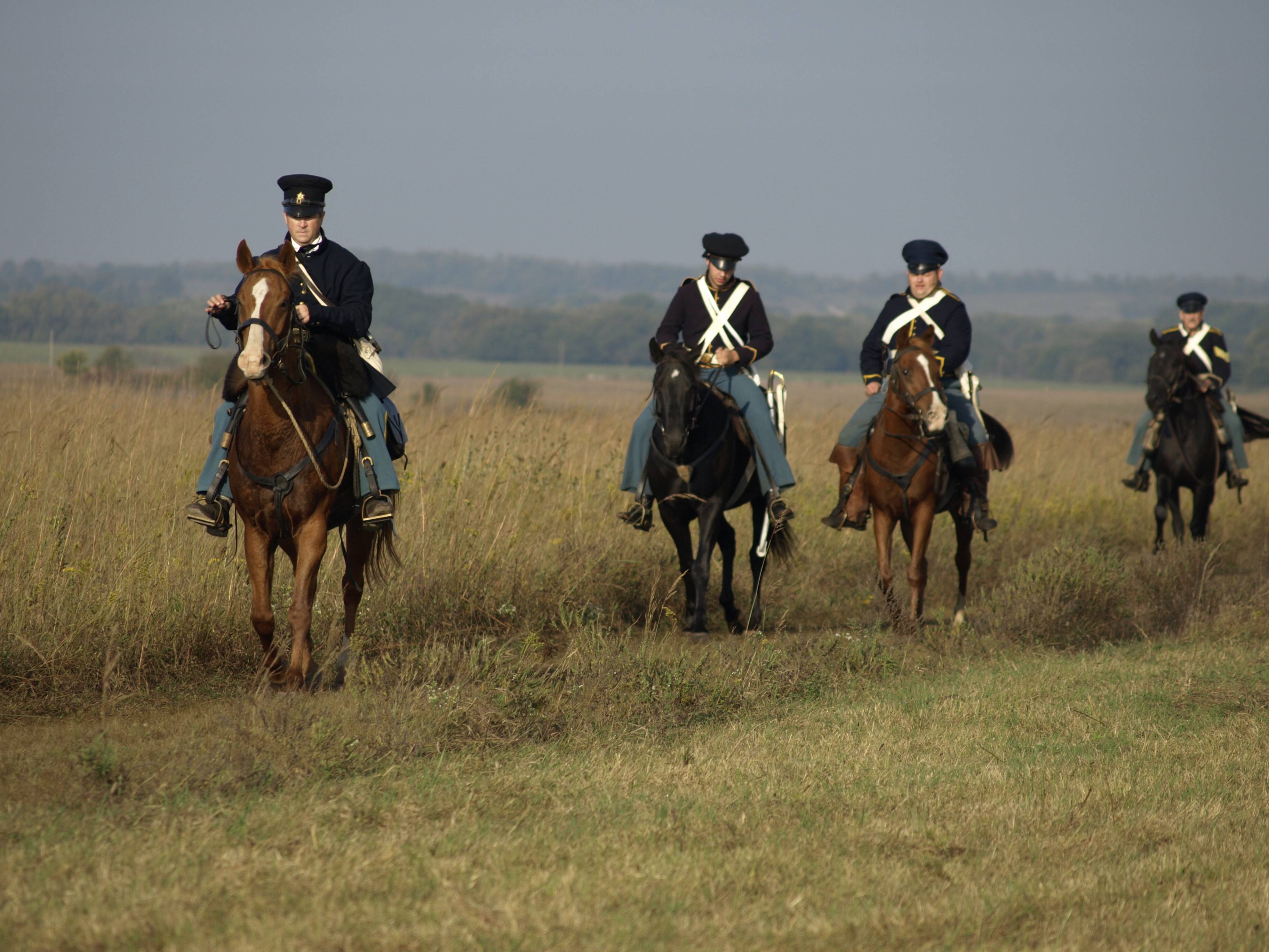 Four soldiers on horseback in a line riding through prairie grass