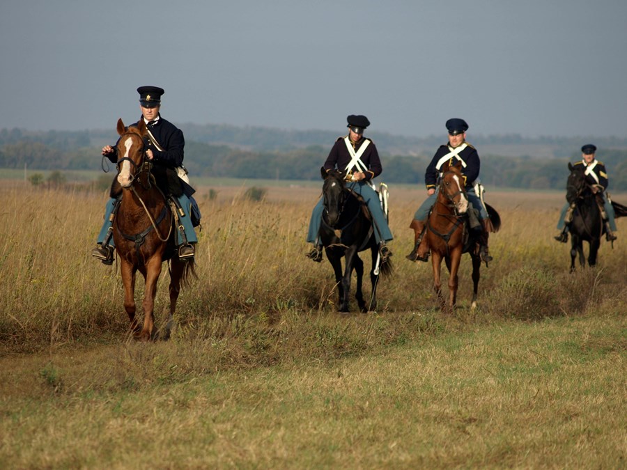 Four soldiers on horseback in a line riding through prairie grass.