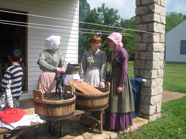 Women dressed as laundresses with laundry buckets and scrub boards stand on a stone porch.