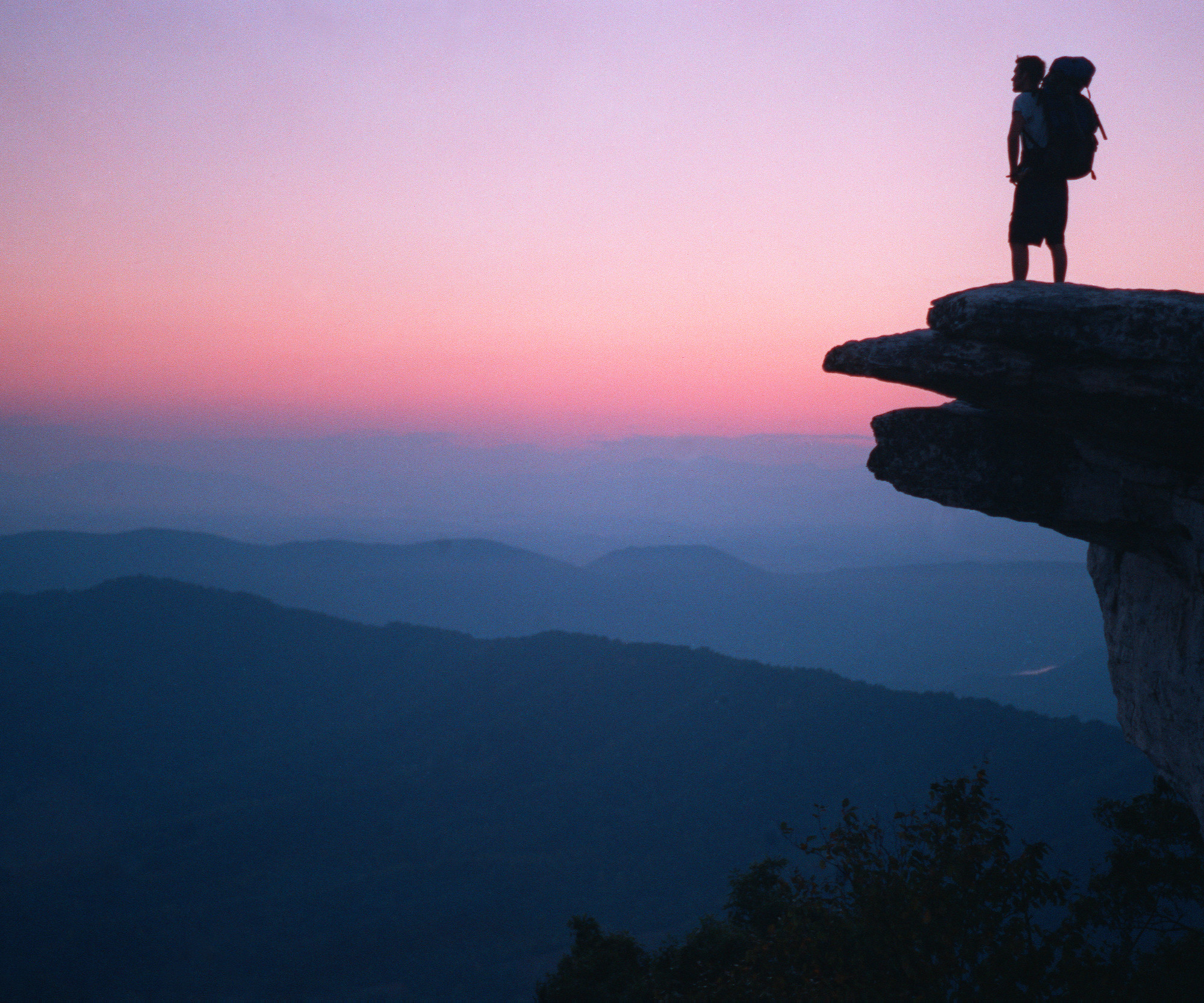 Silhouette of a man with backpack standing on McAfee Knob at sunset with mountains in the distance.