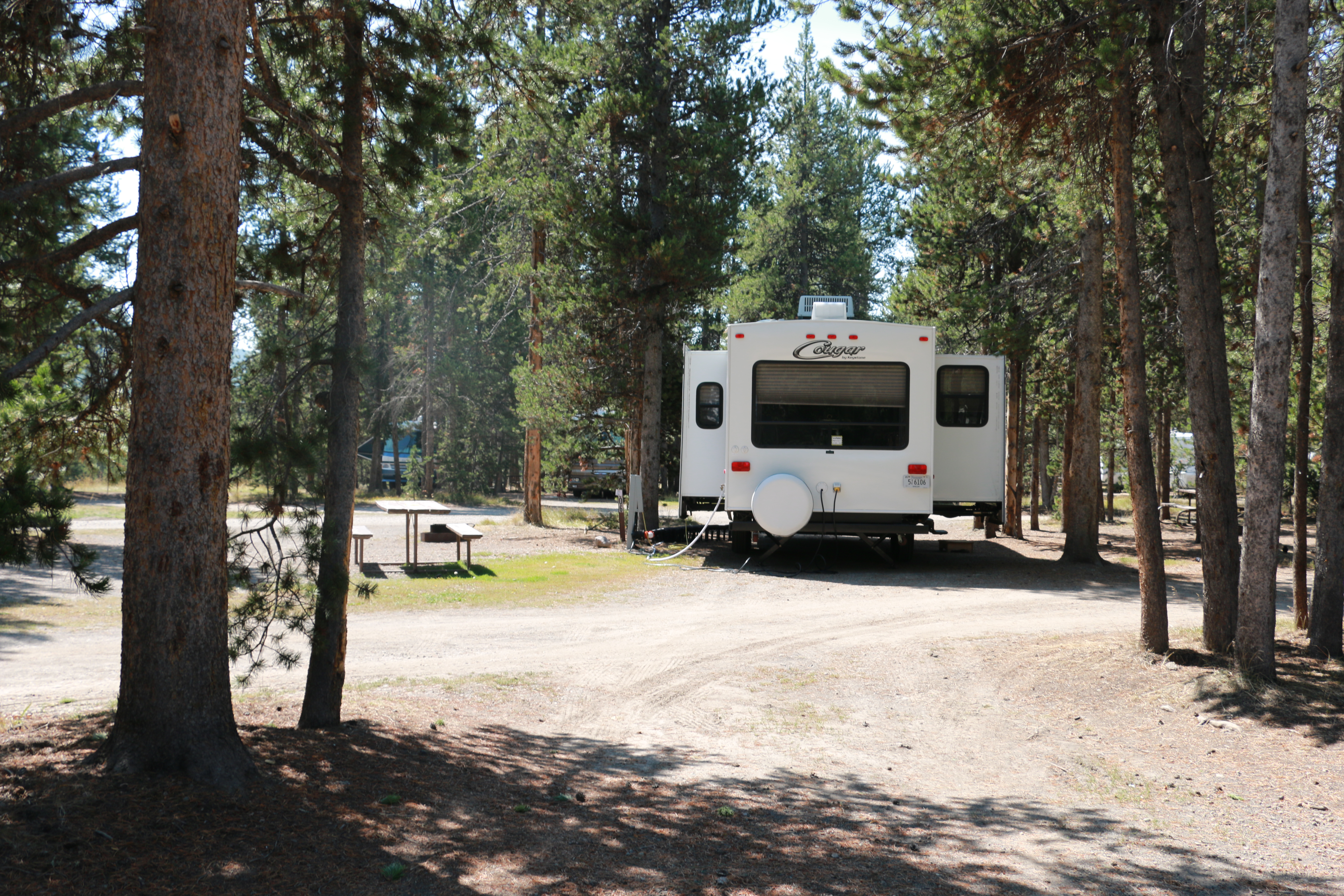white camper trailer with slideouts extended and picnic table partly shaded by conifers