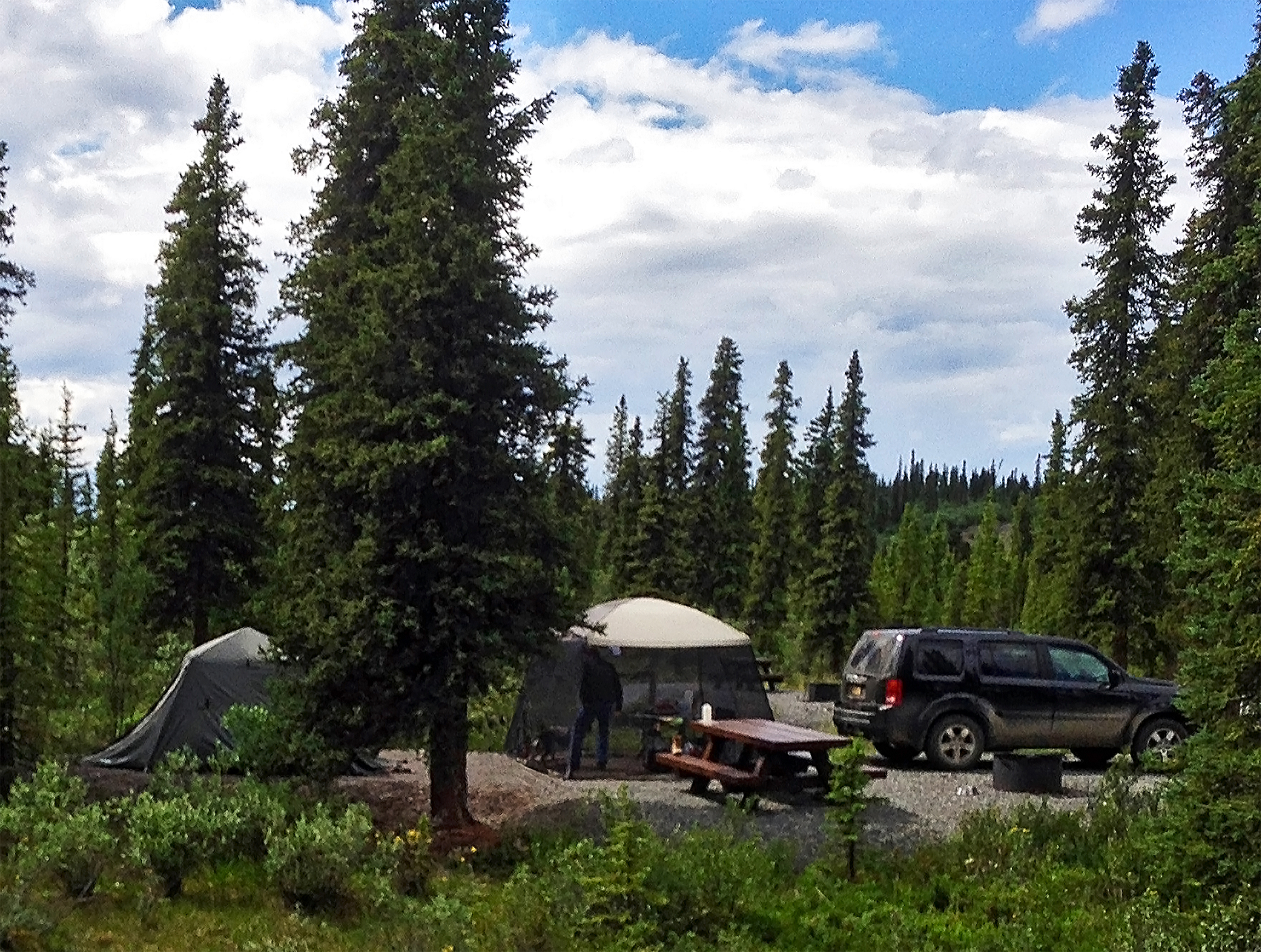 Campsite in the forest with tents, picnic table, and vehicle.