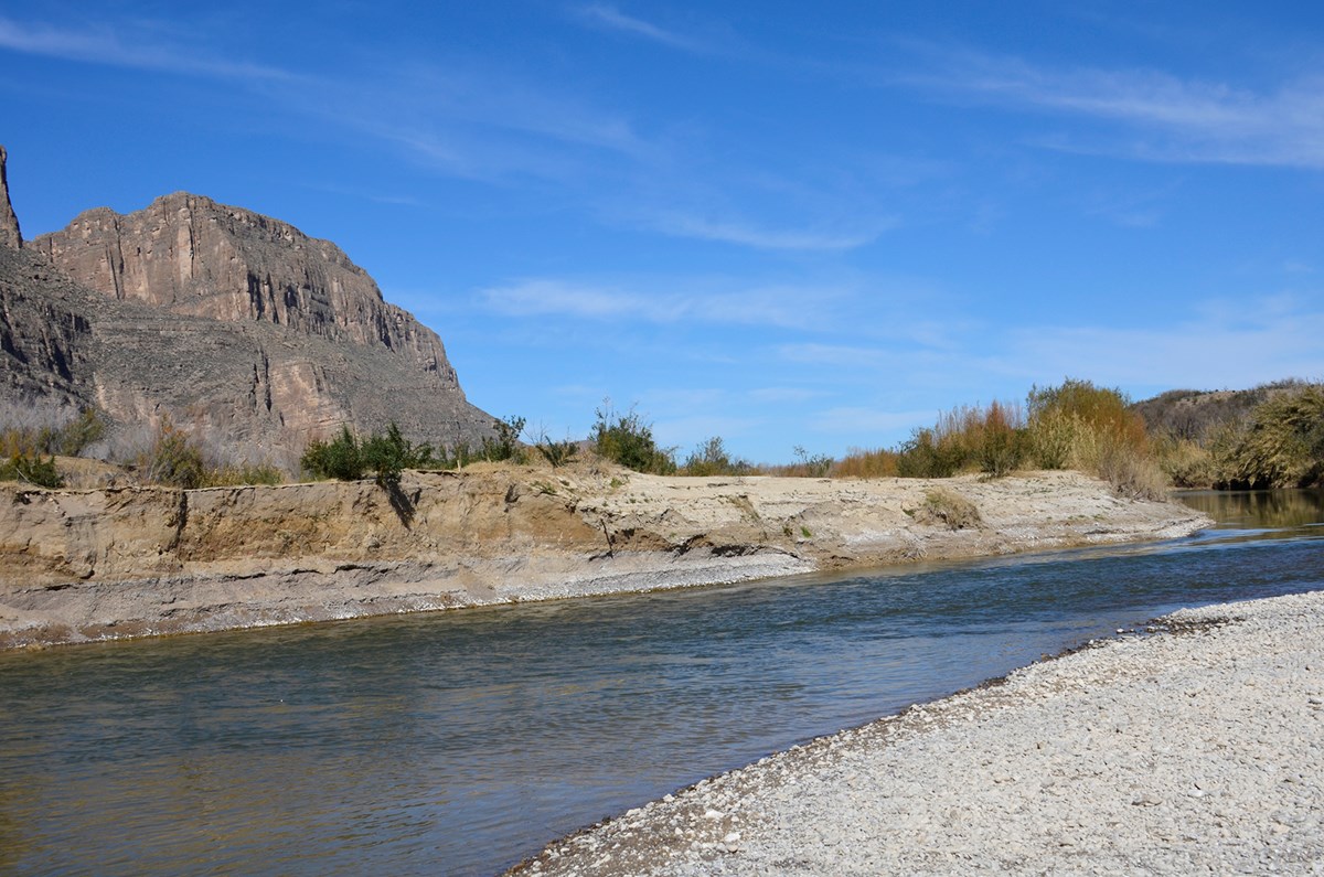 A desert river with a rugged landscape of cliff faces and hills in the background.