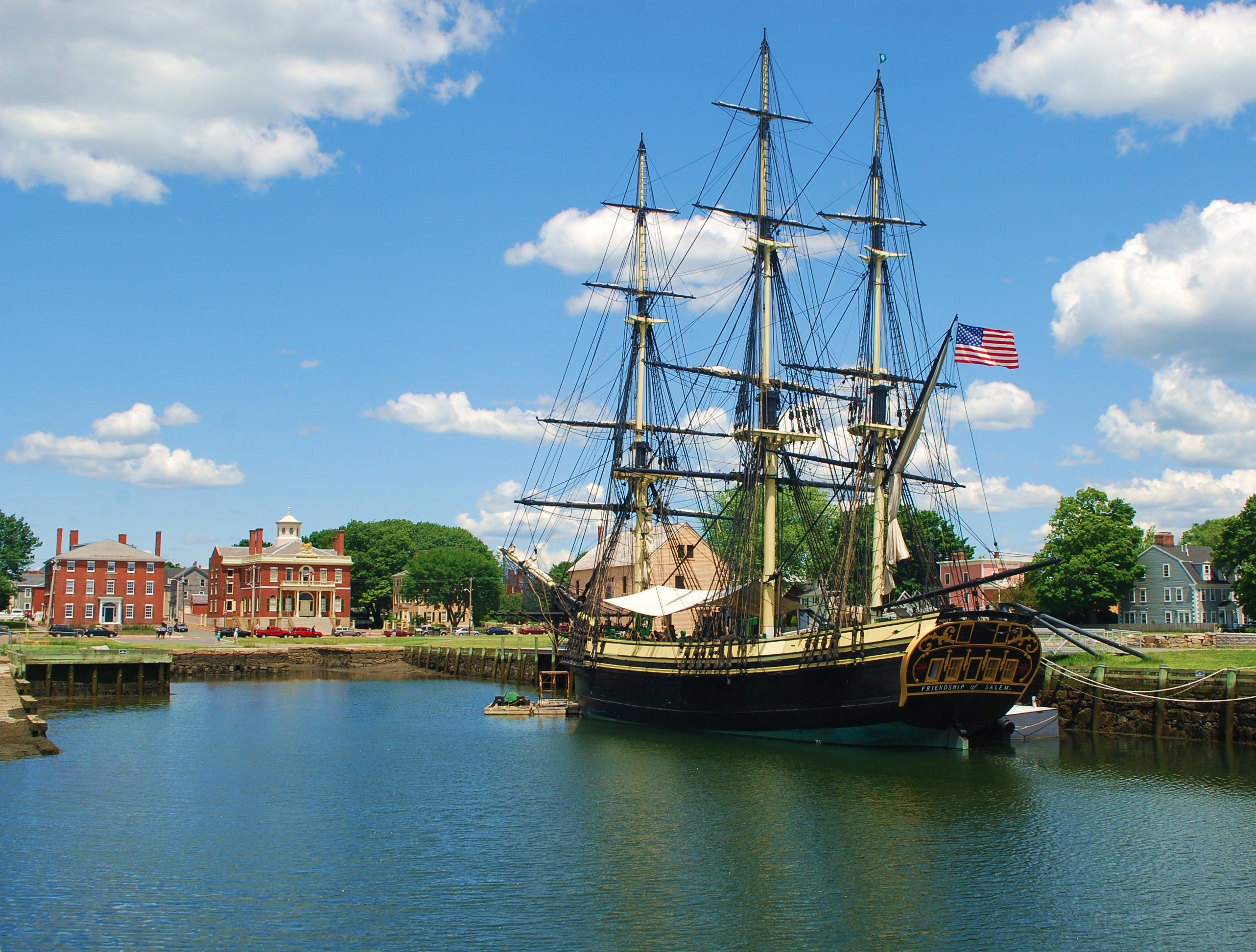 A three mast tall ship on the water under a blue sky with red brick buildings on the shore.