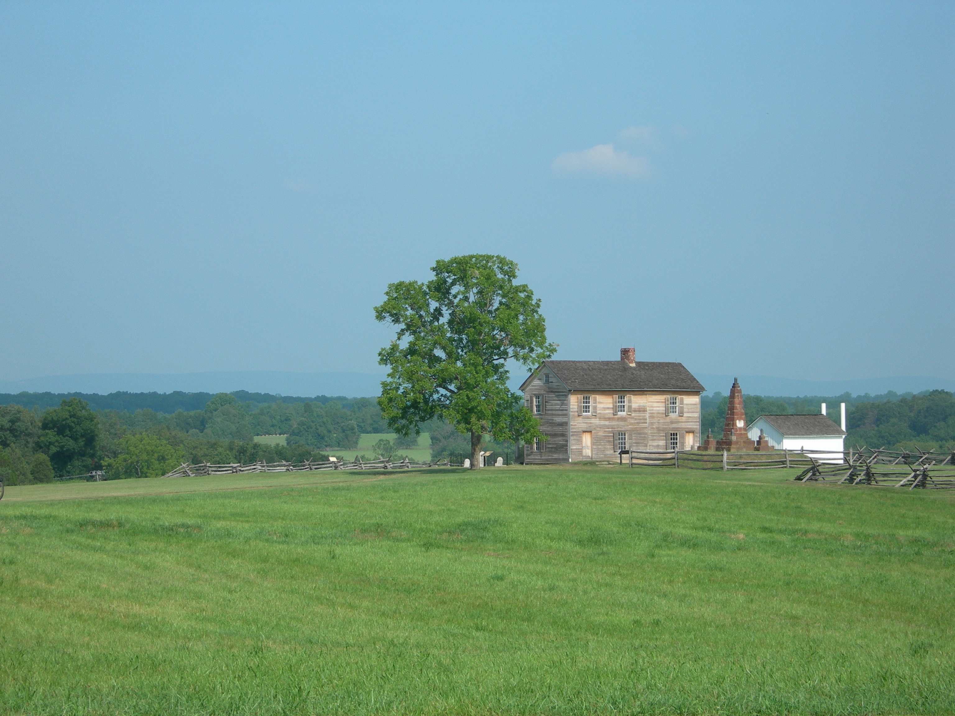 Rustic farmhouse, flanked by shade tree, stone monument, and white outbuilding, on top of a hill.