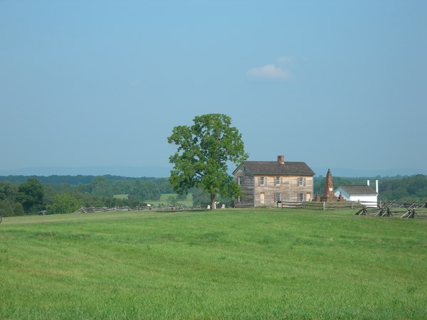 Rustic farmhouse, flanked by shade tree, stone monument, and white outbuilding, on top of a hill.