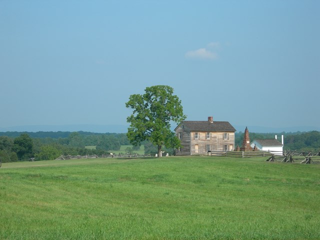 Rustic farmhouse, flanked by shade tree, stone monument, and white outbuilding, on top of a hill.
