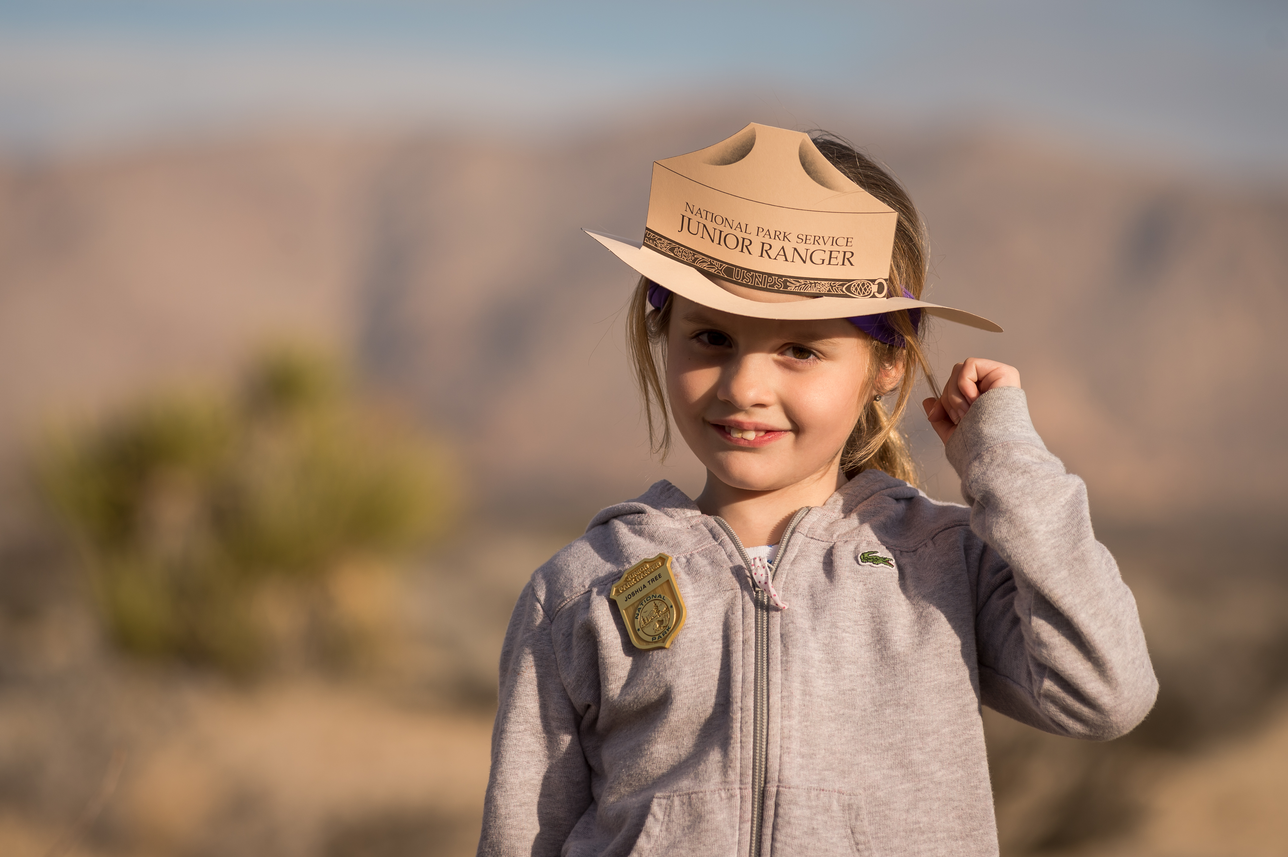 a little girl smiles while wearing a Jr. Ranger hat and badge