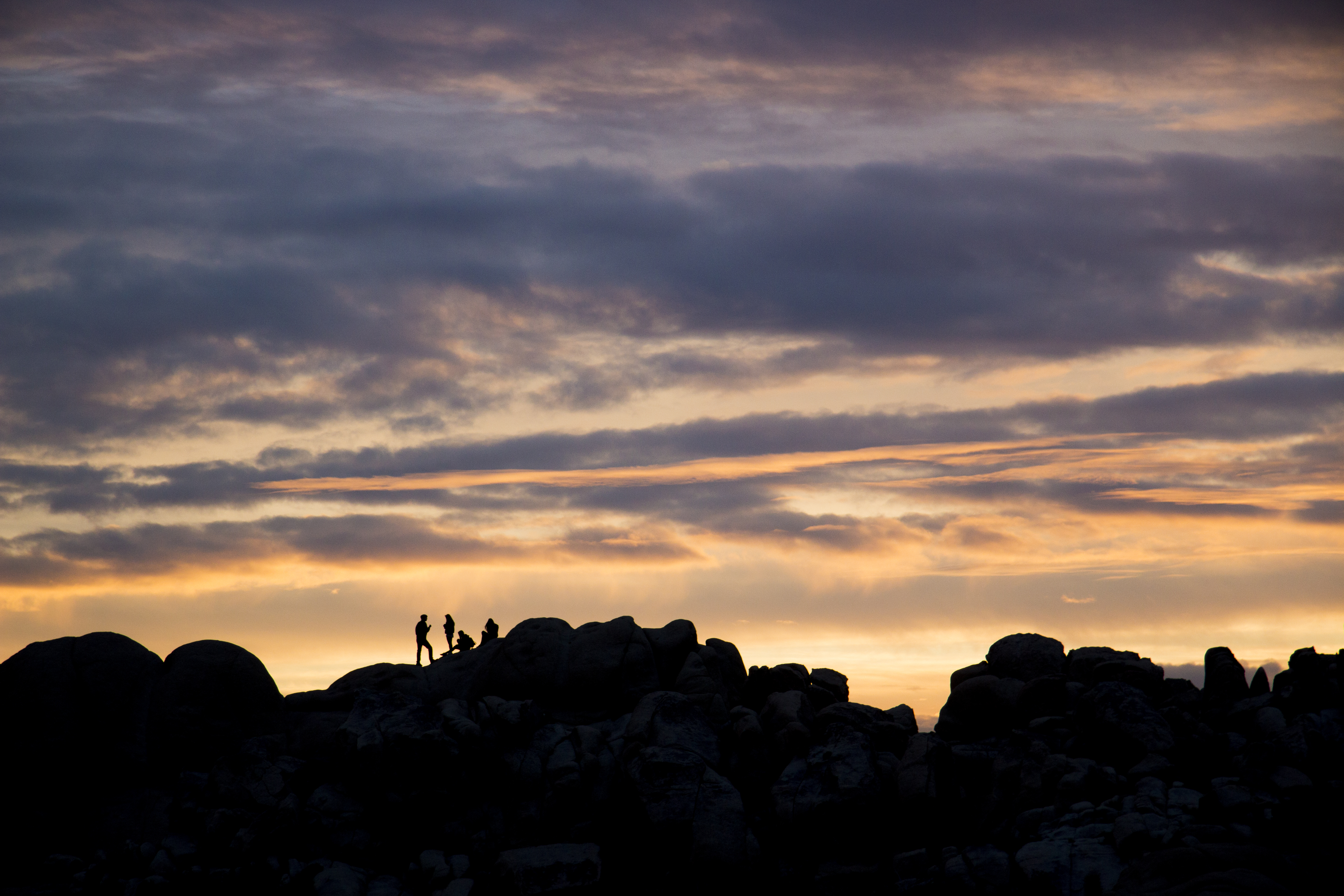 people climbing on boulders are silhouetted against a colorful sunset sky