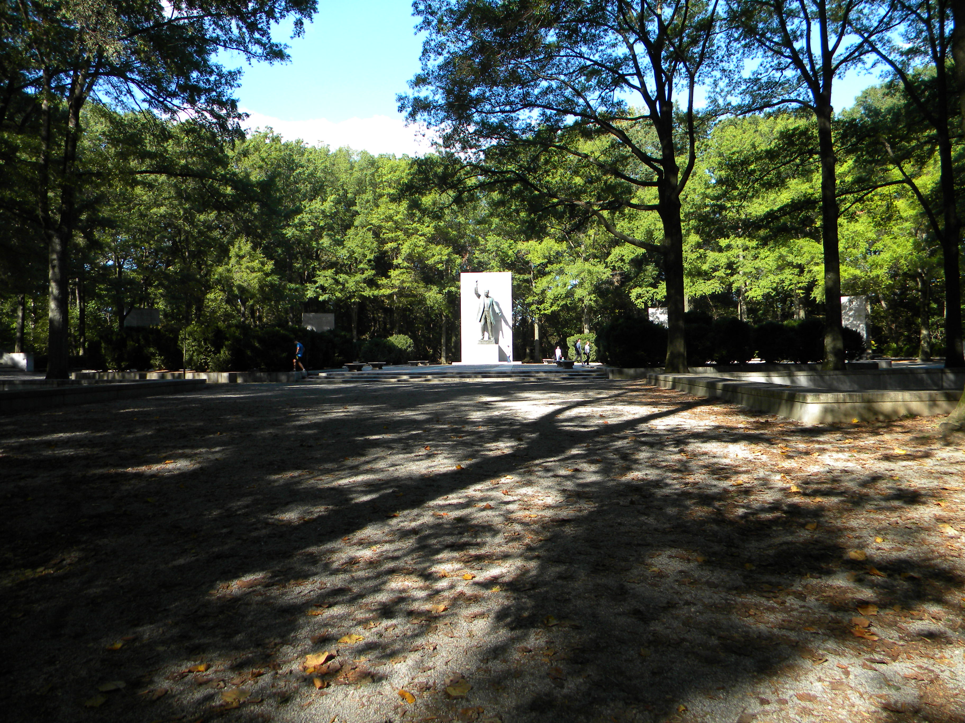 long stone plaza with statue of Theodore Roosevelt at end surround by the shadows of tall trees