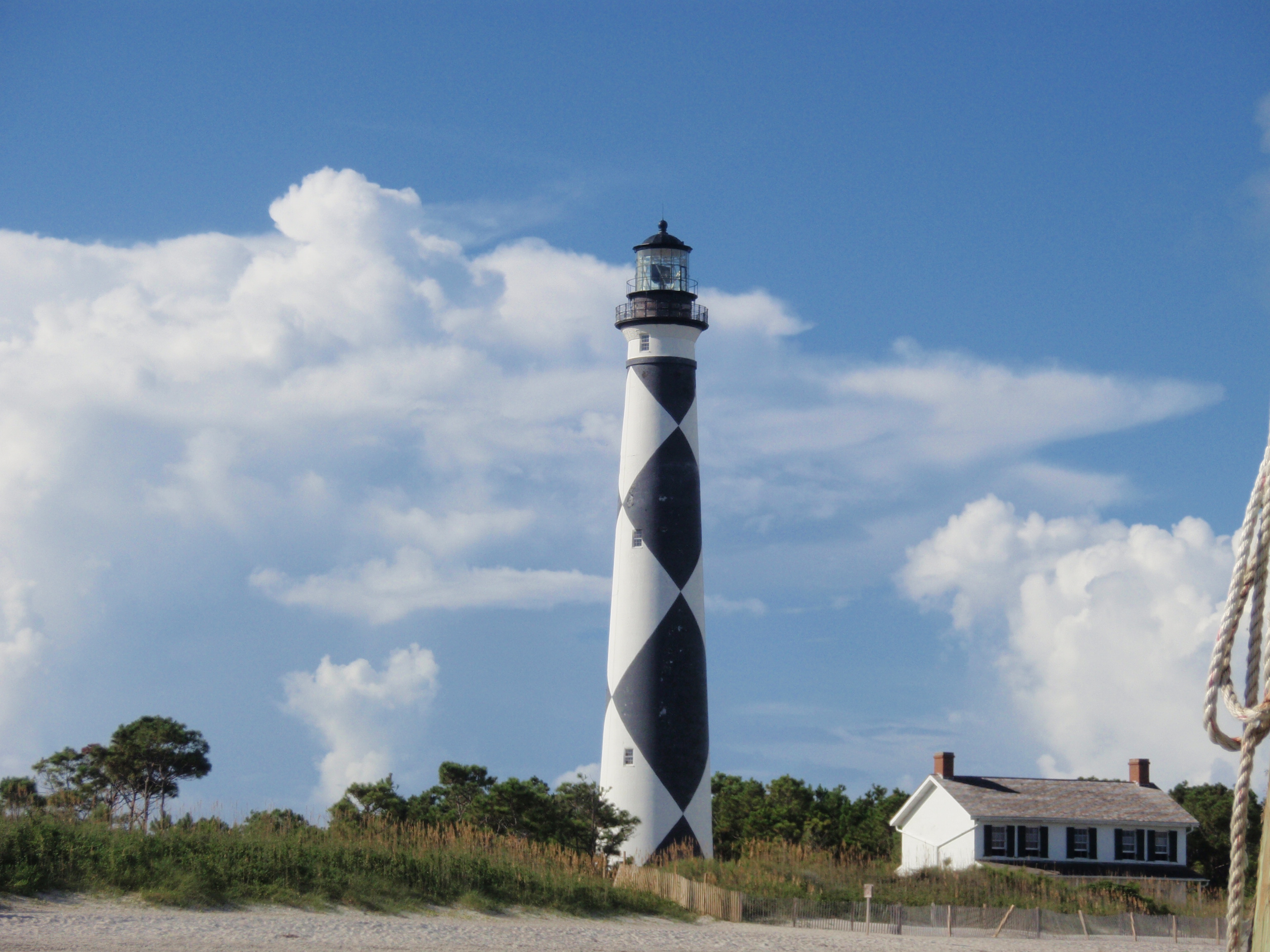 : Black & white patterned tower of the Cape Lookout Lighthouse stands against a blue & white cloud f