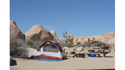 A tent, fire pit, picnic table are in a campsite surrounded by boulders.