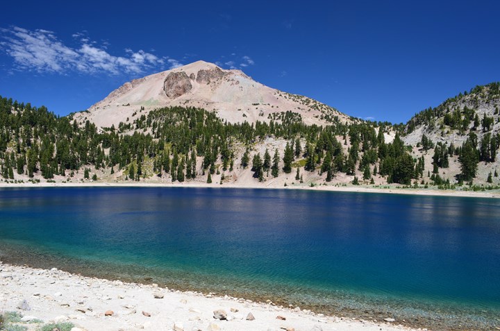 Lassen Peak rising above a tree-lined, deep blue Lake Helen under a bright sky