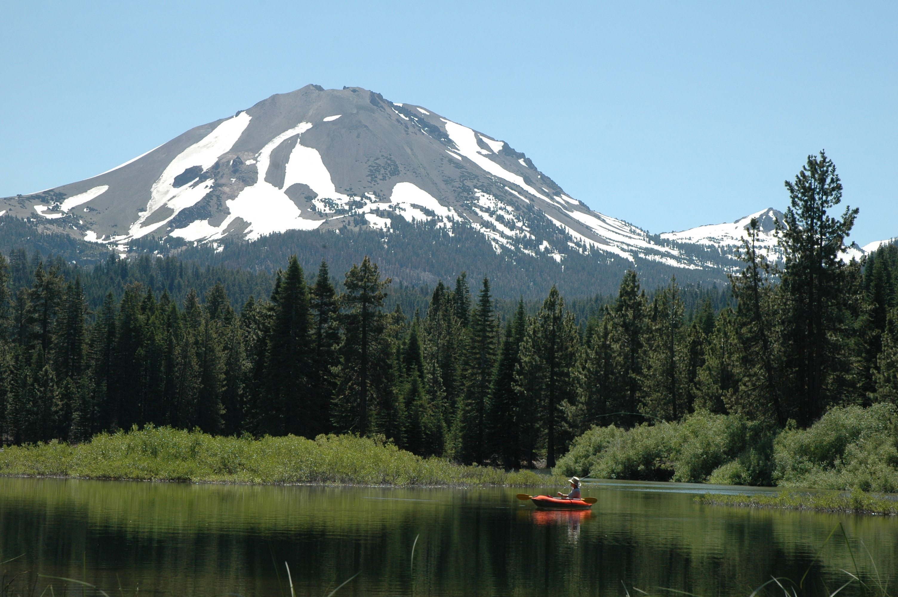 A fisherman casts a line from a boat below a snow-dotted volcanic peak.