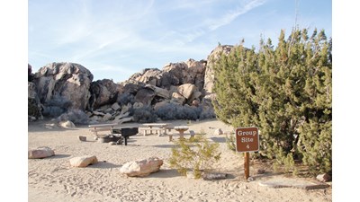A sign that reads "Group Site 4". Behind are three picnic tables, a fire ring and a fire grate.