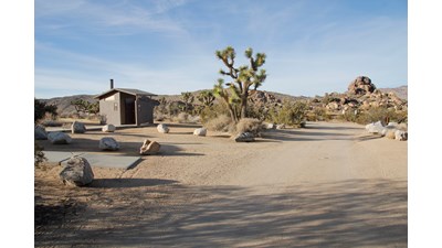 An dirt road and a small paved walking path lead to a vault toilet.